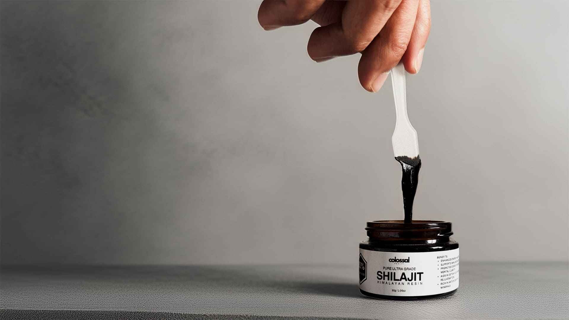 Hand using a spoon to scoop shilajit from a jar labeled 'Colossal Health Shilajit' on a grey background