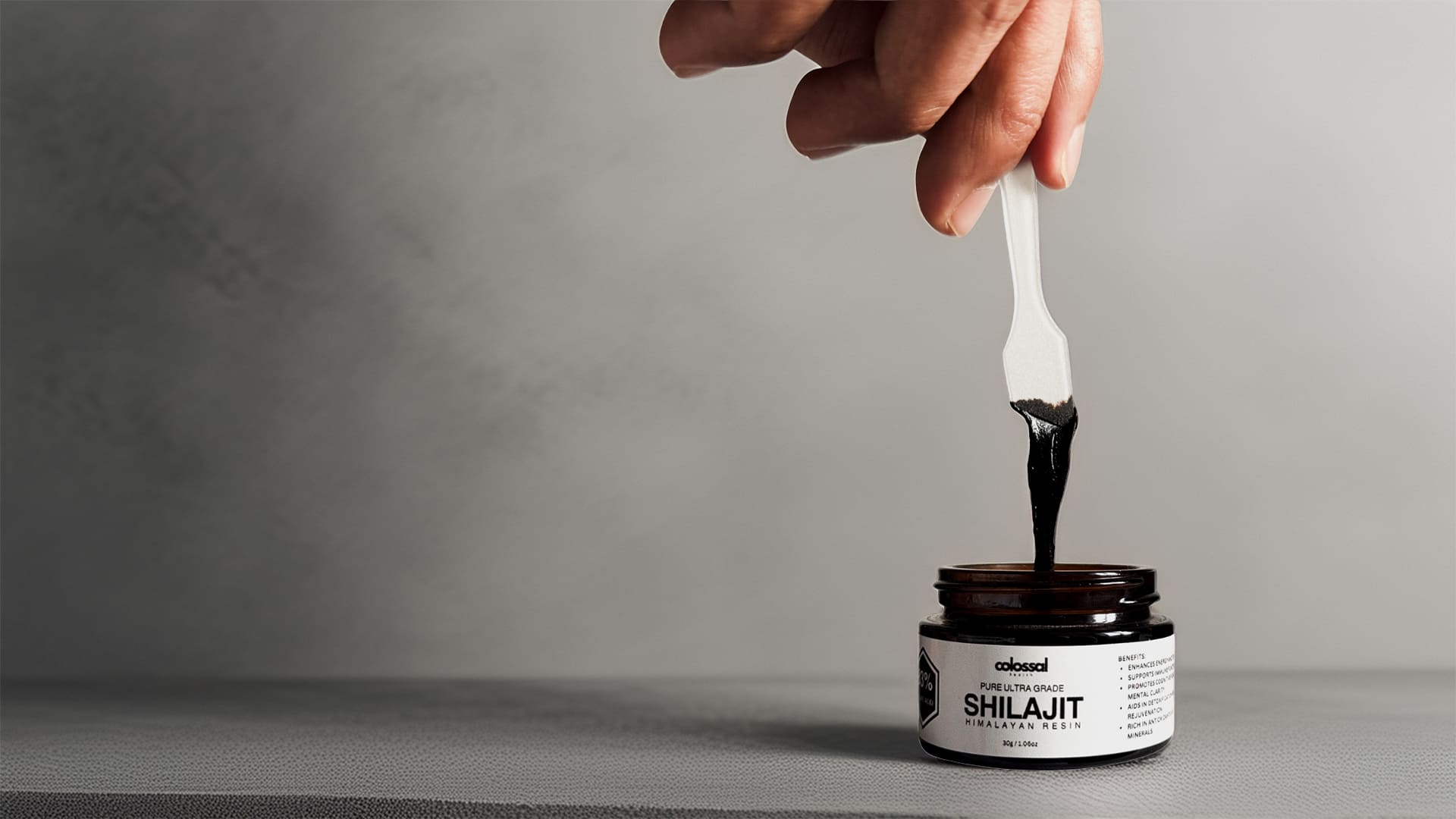 Hand using a spoon to scoop shilajit from a jar labeled 'Colossal Health Shilajit' on a grey background