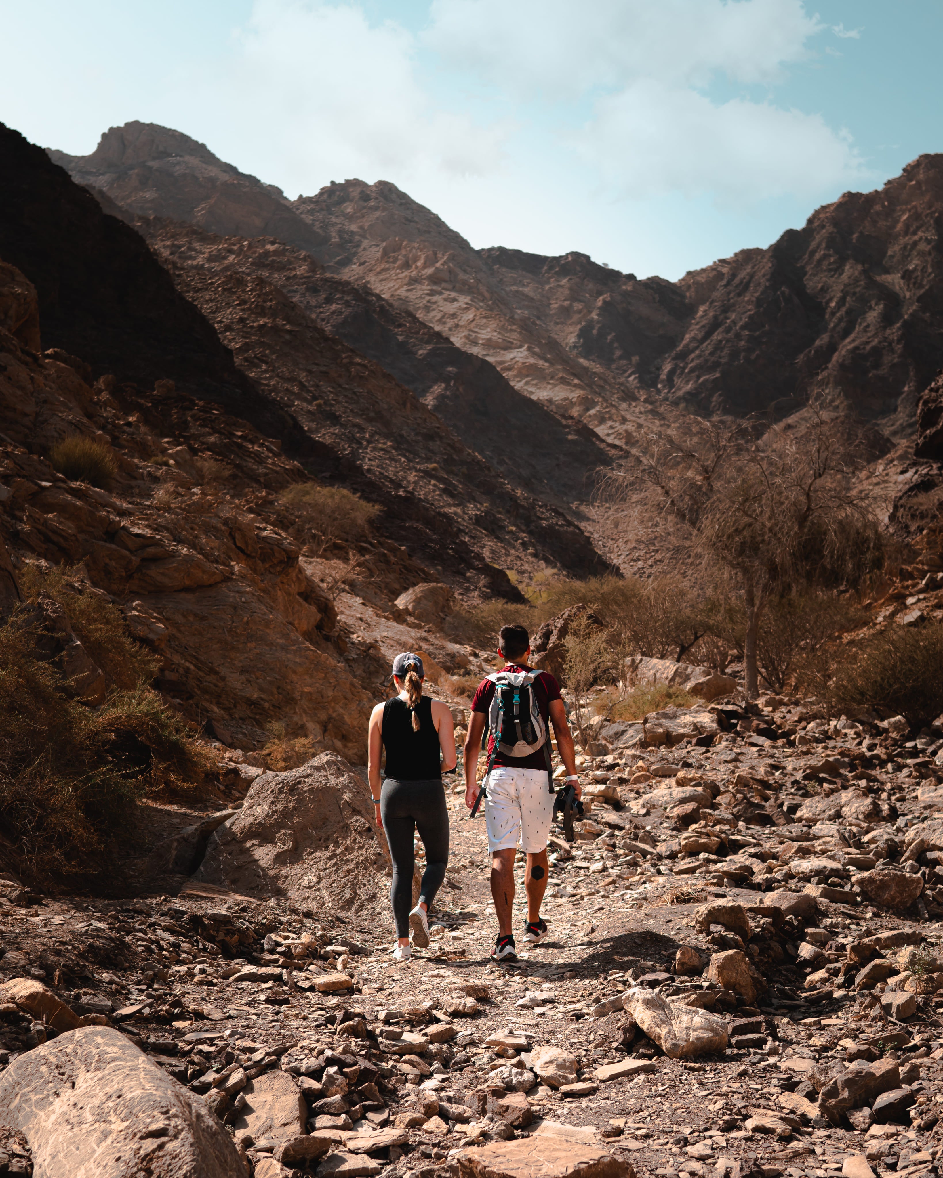 Two hikers walking on a rocky trail in a mountainous desert landscape.